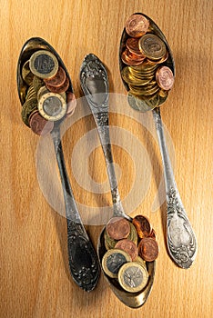 Old spoons and euro and eurocent coins lie on the surface of a wooden table