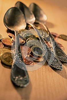 Old spoons and euro and eurocent coins lie on the surface of a wooden table