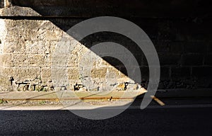 Old stone wall of an underpass divided in two by the shadow of the bridge. Weathered sidewalk and road in front.