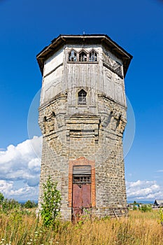 Old stone water tower with windows and a wooden roof against the background of blue sky and green trees on a sunny summer day