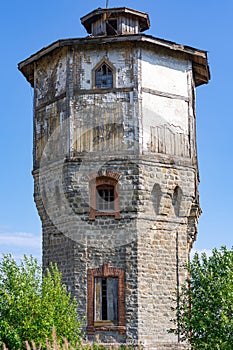 Old stone water tower with windows and a wooden roof against the background of blue sky and green trees on a sunny summer day