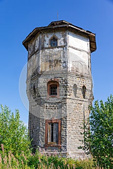 Old stone water tower with windows and a wooden roof against the background of blue sky and green trees on a sunny summer day.