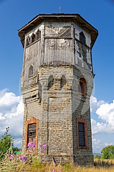 Old stone water tower with windows and a wooden roof against the background of blue sky and green trees on a sunny summer day