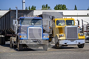 Old style day cab big rig semi trucks with dump trailers standing in row on the marked spots warehouse parking lot