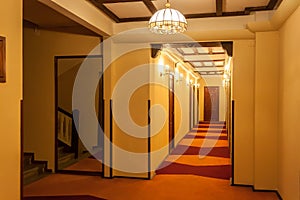 Old style hotel hallway with wooden brown doors, reddish carpet, and yellow walls. Mirror seen near the floor stairs