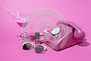 Old style rotary telephone on pink background with cocktail, sunglasses and nail polish