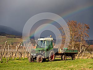 An old tractor stands in front of vineyards amid a rainbow and the Vosges mountains in Alsace. Bright spring landscape. Awakening