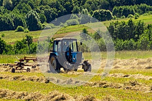 An old tractor turns over the mowed hay on a Sunny summer morning for better drying. Fodder for cows for the winter