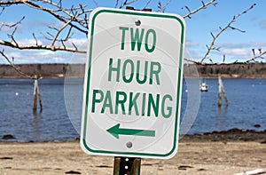 Old two hour parking sign with a left arrow at an angle in front of a beach and ocean in the springtime in New England