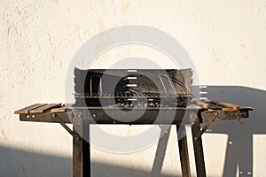 An old used charcoal grill stands in the sun in front of a light wall. The coal is still in the grill. Grabbers are lying next to