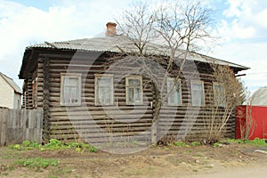 An old village wooden log house with a slate roof and a brick chimney. The tree grows in front of the house