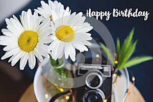 Old vintage camera with a bouquet of daisy flowers on a wooden board. Inscription Happy Birthday