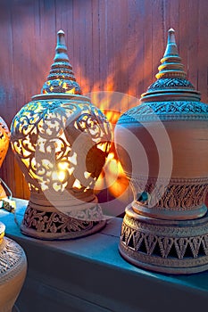 The old vintage clay carving electric lamp in Thai style with illuminated lighting and pottery on wooden shelf display for sale