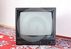 An old vintage TV sits on a bright Soviet carpet on the floor from the 1980s, 1990s, 2000s next to it.