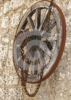 Old vintage wooden wheel with rusty metal chain hanging on a stone house wall. Side view