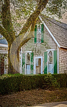 Old weathered vintage wooden shingled house with arched church-like windows and green shutters stand behind tree and hedge