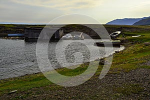 Old white rusty iron boats on green grassy shore of small round bay, with barn on the water. Blue mountains