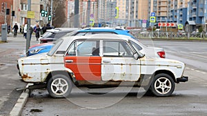 An old white rusty Soviet car with a red door is parked on the street