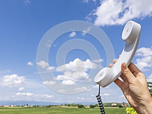 An old white telephone handset is lifted by one hand towards a beautiful blue sky with white clouds and the countryside