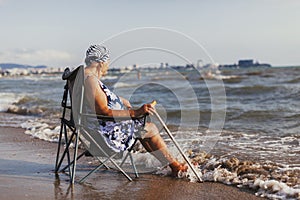 Old white woman in a swimsuit and a bandana sits in a tourist chair near the sea