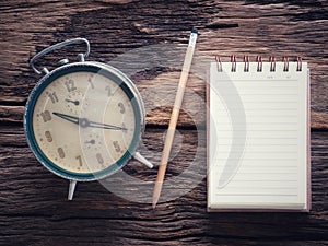 Old wind clock with notebook and pencil on wood table in vintage color scheme