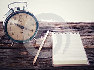 Old wind clock with notebook and pencil on wood table in vintage color scheme