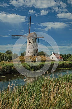 Old windmill next to canal with bushes and grove in the background in the late afternoon light and blue sky, near Damme.