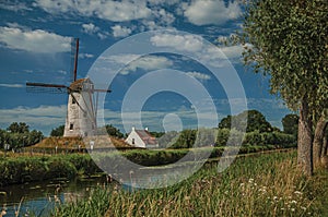 Old windmill next to canal with bushes and grove in the background in the late afternoon light and blue sky, near Damme.