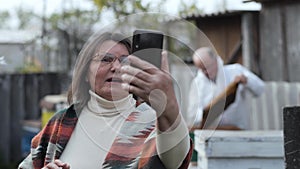 Old woman blogger with smartphone in her hands speaks via video link about beekeeping showing a man in a protective suit