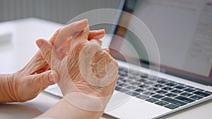 Old woman company manager holds wrinkly hands over keyboard and types against blurred table with light reflections at home