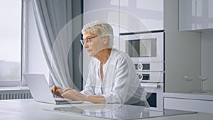 Old woman company manager types on grey laptop sitting at large white table in kitchen against window with curtains