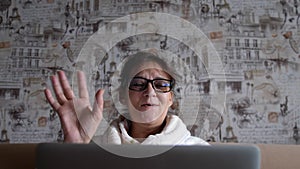 Old woman in glasses having a video call on the laptop smiling and talking happily indoors in a cozy apartment. doctor