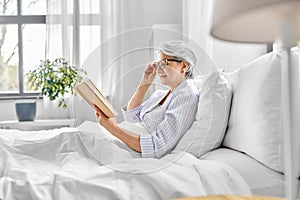 old woman in glasses reading book in bed at home