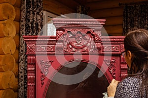 Old woman gluing carved wood ornaments to wooden cardboard of light red color with glass in front in wooden cottage