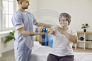 Old woman at medical clinic trains with physiotherapist and does exercises using dumbbells.