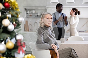 Old woman sitting at the kitchen table with her back to couple quarreling together in the kitchen with Xmas-tree