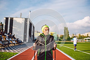 Old woman in sportswear practicing nordic walking outdoors on rubber treadmill in stadium. Older female walk by scandinavian walk