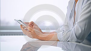 Old woman in white blouse wrinkly hands type on smartphone over shiny table with light reflections against window