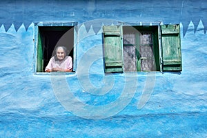 Old woman at the window in old traditional blue house