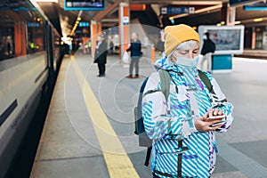 Old woman in yellow hat waiting on station platform with backpack on background train using smart phone. Railroad