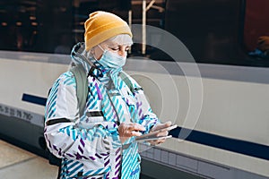 Old woman in yellow hat waiting on station platform with backpack on background train using smart phone. Railroad