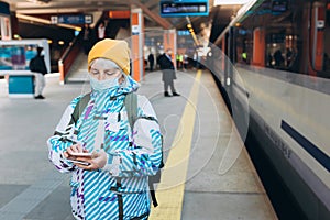 Old woman in yellow hat waiting on station platform with backpack on background train using smart phone. Railroad