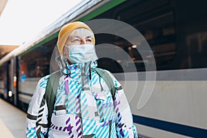 Old woman in yellow hat waiting on station platform with backpack on train background. Railroad transport concept