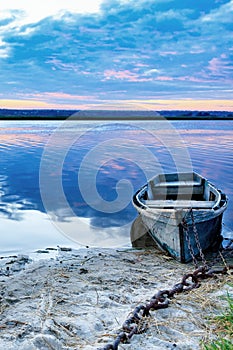 An old wooden boat is moored to the bank with a rusted iron chain on the bank of a calm river against the morning horizon