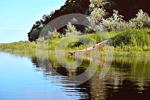 old wooden boat on the river bank against the background of green grass and bushes, reflections in the water