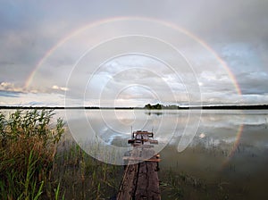 old wooden bridge on the lake on the background of a beautiful rainbow in a cloudy sky. Landscapes of the hinterlands of