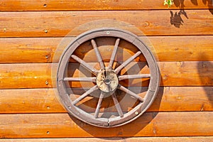 An old wooden carriage wheel with metal fittings hangs on the wall of a wooden house for decoration. Wooden spoke wheel. Outdoor