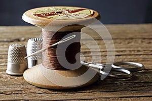 OLD WOODEN COTTON REEL WITH STRONG BROWN COTTON THREAD, A NEEDLE AND THIMBLES AND A PAIR OF SCISSORS IN THE BACKGROUND