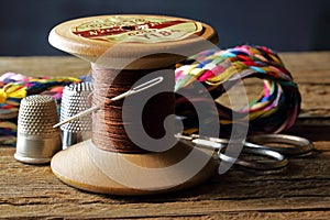 OLD WOODEN COTTON REEL WITH STRONG BROWN COTTON THREAD, A NEEDLE AND THIMBLES, A PAIR OF SCISSORS AND A COLORFUL HTREAD BRAID IN T