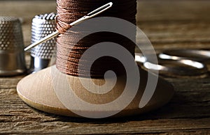 OLD WOODEN COTTON REEL WITH THICK BROWN COTTON THREAD, A NEEDLE AND THIMBLES AND A PAIR OF SCISSORS IN THE BACKGROUND